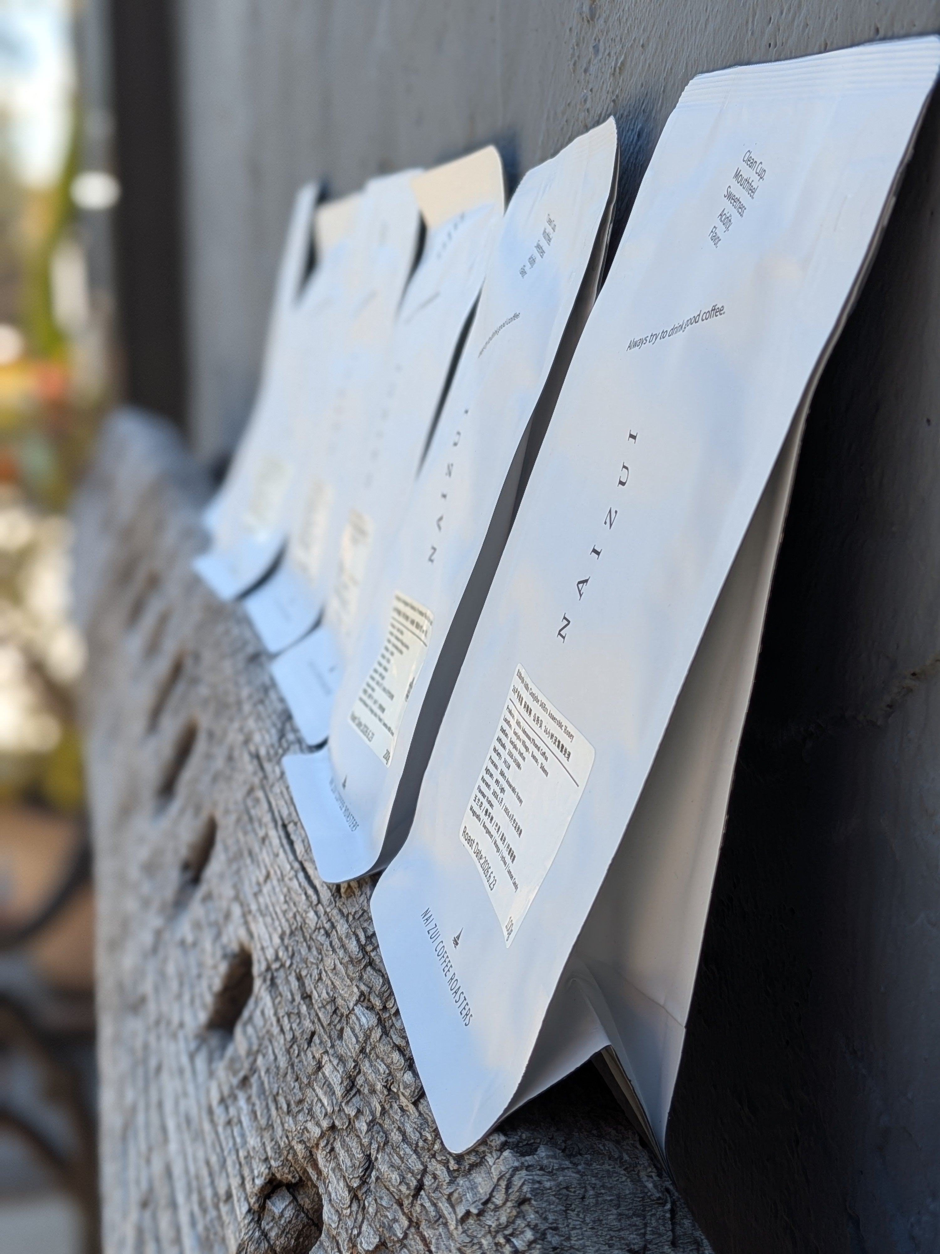 Several white flat pouches of coffee are arranged leaning against a dark wall on a weathered wooden surface. The front pouch is most in focus, with the others softly blurred in the background.