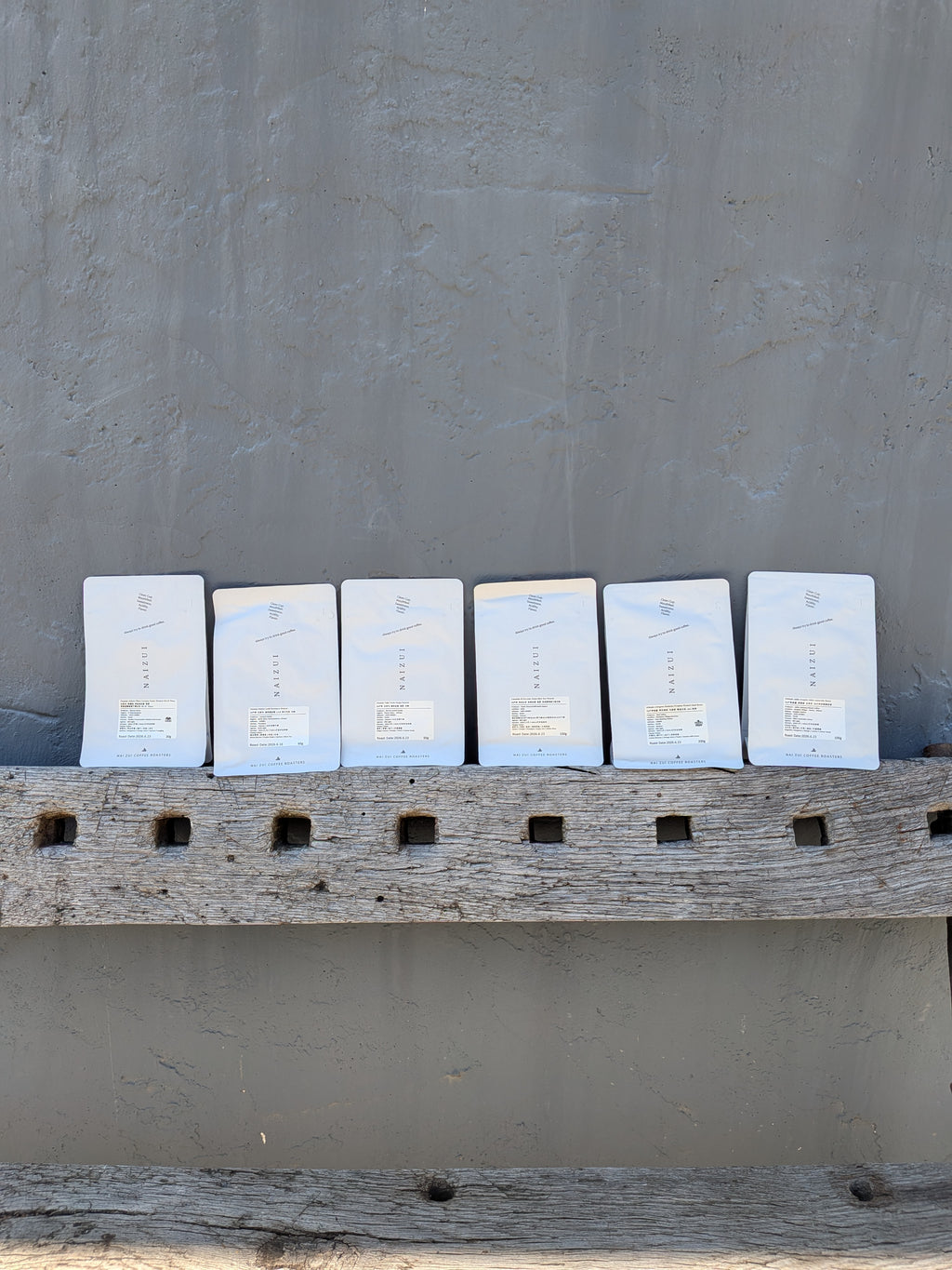 Six white coffee bags arranged in a row on a rustic wooden ledge against a textured gray wall.