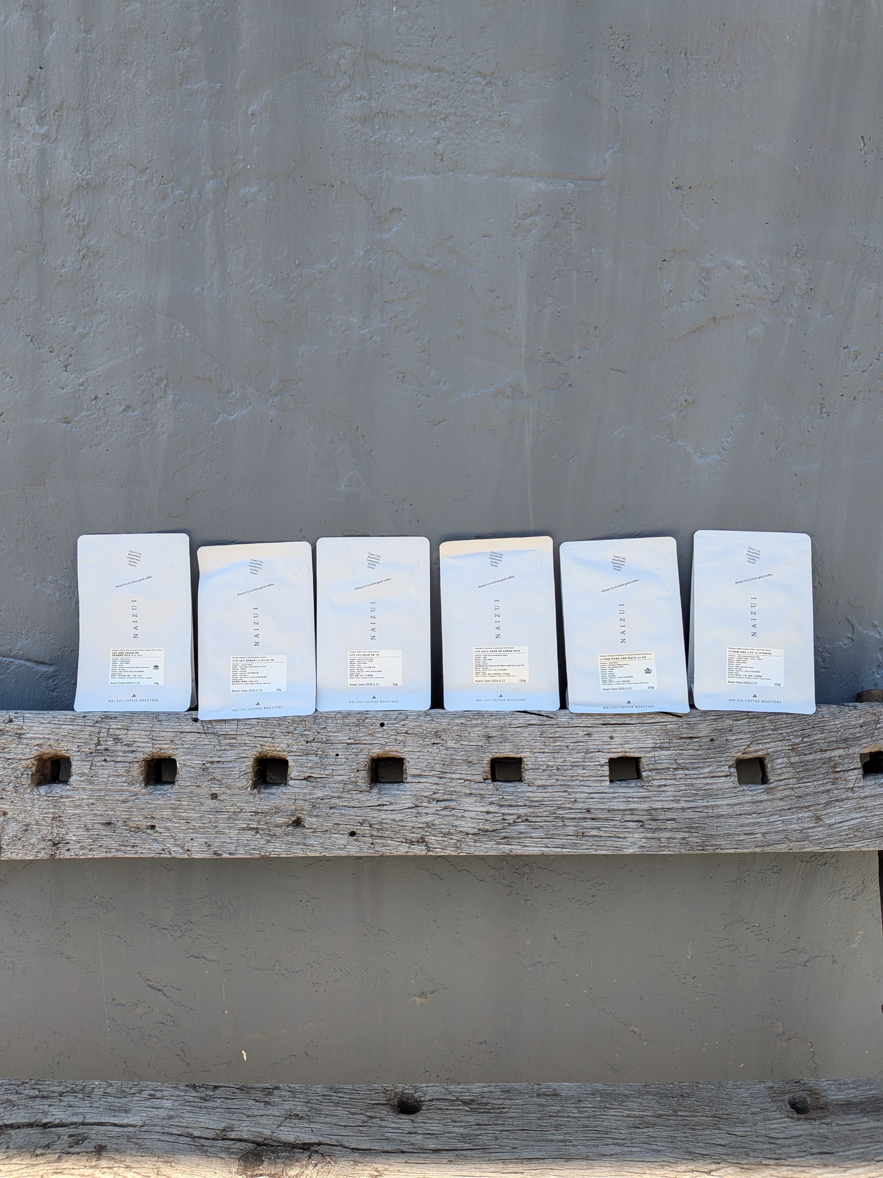 Six white coffee bags arranged in a row on a rustic wooden ledge against a textured gray wall.