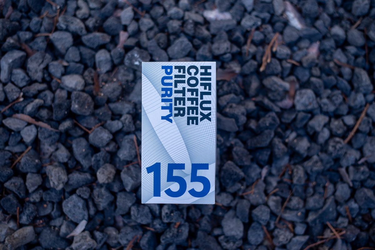 A rectangular HIFLUX Coffee Filter Purify replacement filter card, printed with blue text and the number 155, placed on a bed of dark gray gravel.