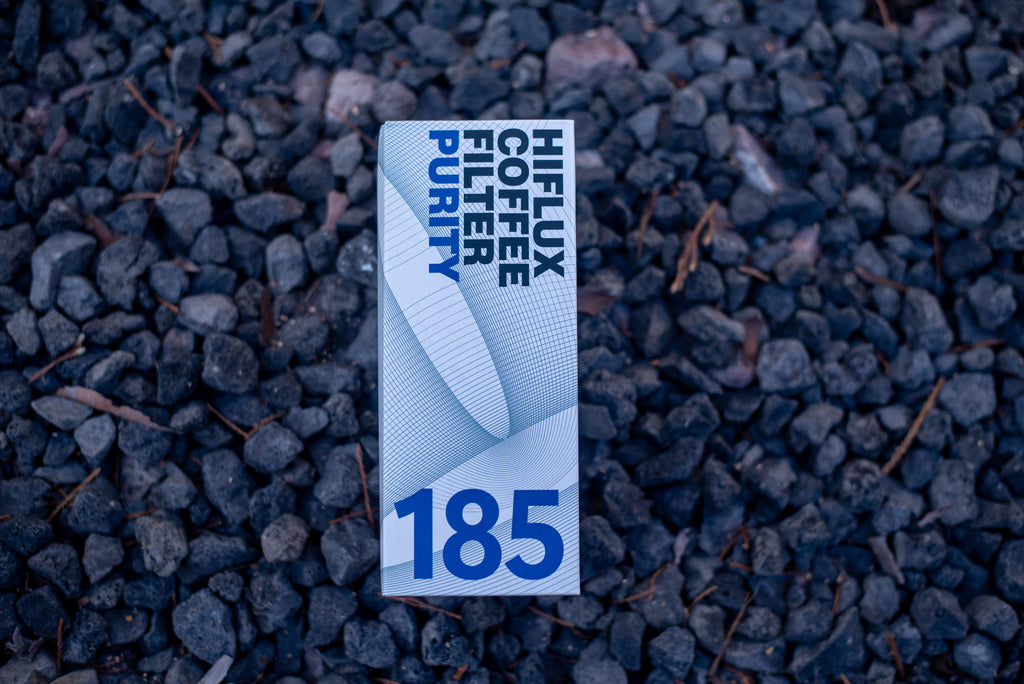 A single rectangular package of coffee stands upright on dark gravel. The package is white with blue text reading 'HIJLUX COFFEE CUTTER PURITY' and a large '185' near the bottom.