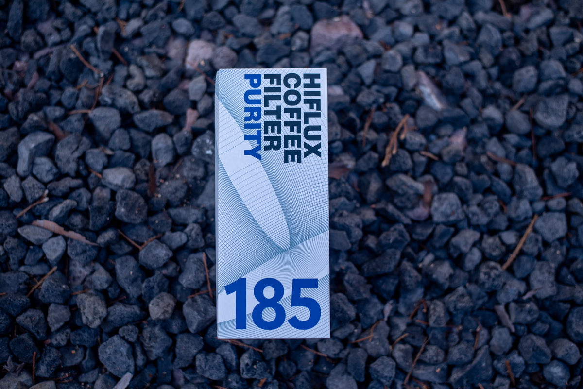 A single rectangular package of coffee stands upright on dark gravel. The package is white with blue text reading 'HIJLUX COFFEE CUTTER PURITY' and a large '185' near the bottom.